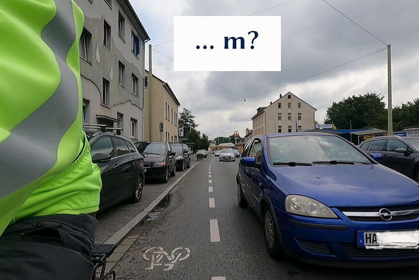 Ein Radfahrer, der auf einem sehr schmalen Schutzstreifen fährt, wird in Kürze von einem Auto überholt, dass nicht von seiner Fahrposition nah an der gestrichtelten Schutzstreifenlinie abweicht.